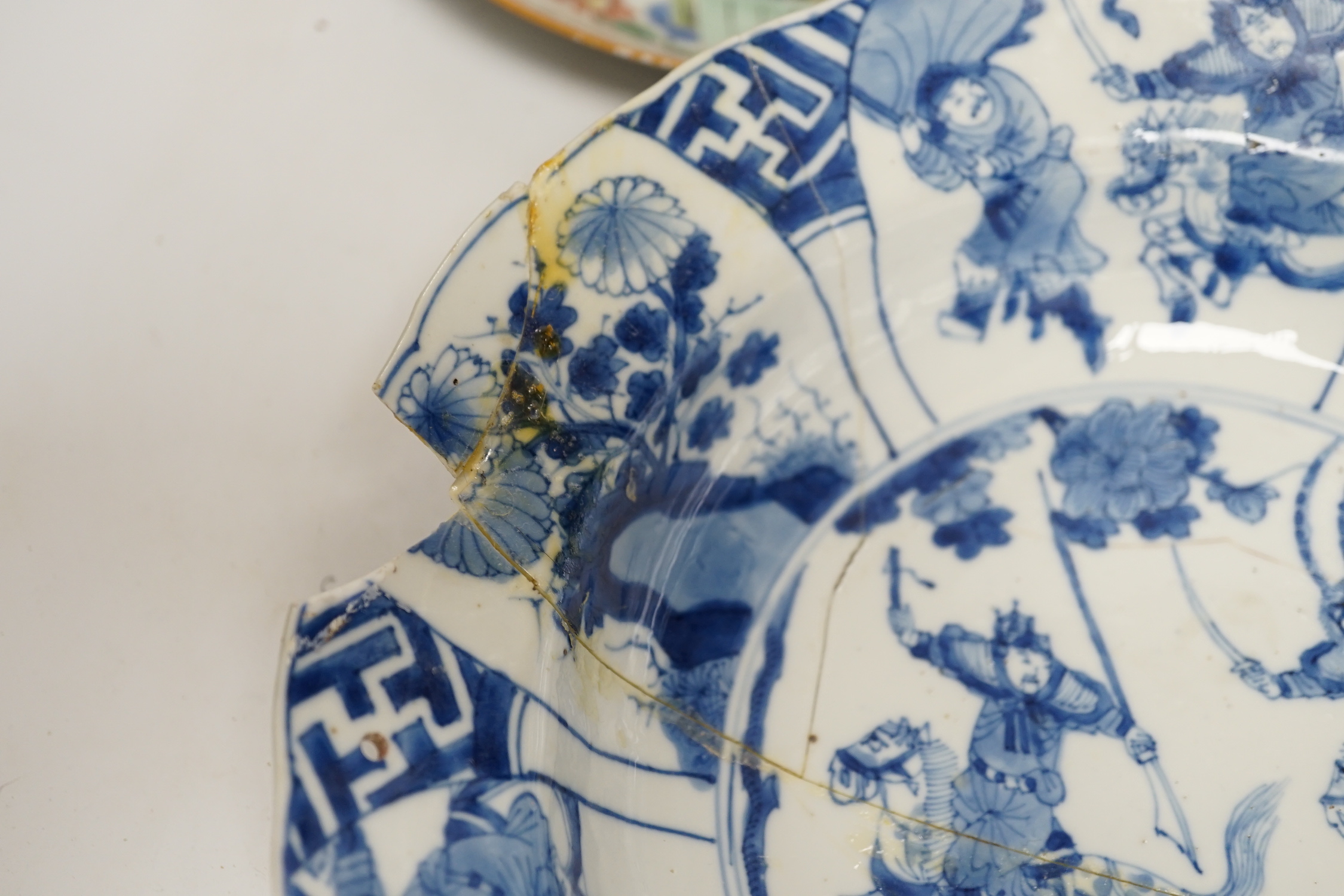 A Chinese blue and white ‘warriors’ bowl, Kangxi mark and period, a similar ‘hunting’ saucer dish, and a famille verte saucer dish, all Kangxi period (3), largest 27cm in diameter